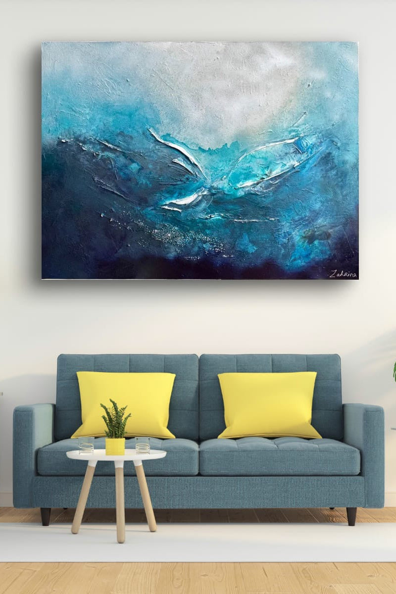 A large landscape abstract canvas print featuring shades of blue, teal, and white, displayed on a white wall above a modern blue sofa with yellow pillows in a styled living room.