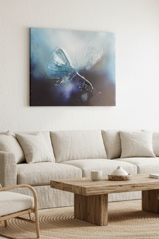 A modern living room featuring a large abstract canvas print with blue, purple, and white tones hanging on the wall above a light beige sofa. The room is styled with a wooden coffee table, woven chairs, potted plants, and a large window looking out to the ocean.