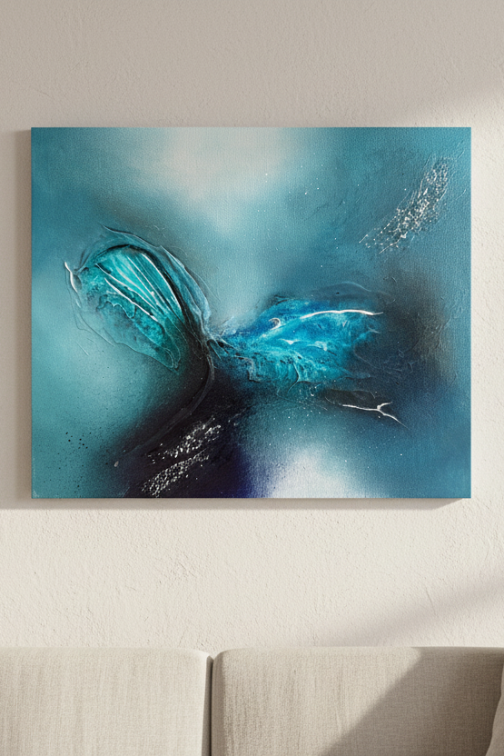 An abstract canvas wall art print featuring vibrant blues and teals with dark accents, displayed on a beige wall in a modern living room setting with a sofa, potted plant, and window overlooking the ocean.